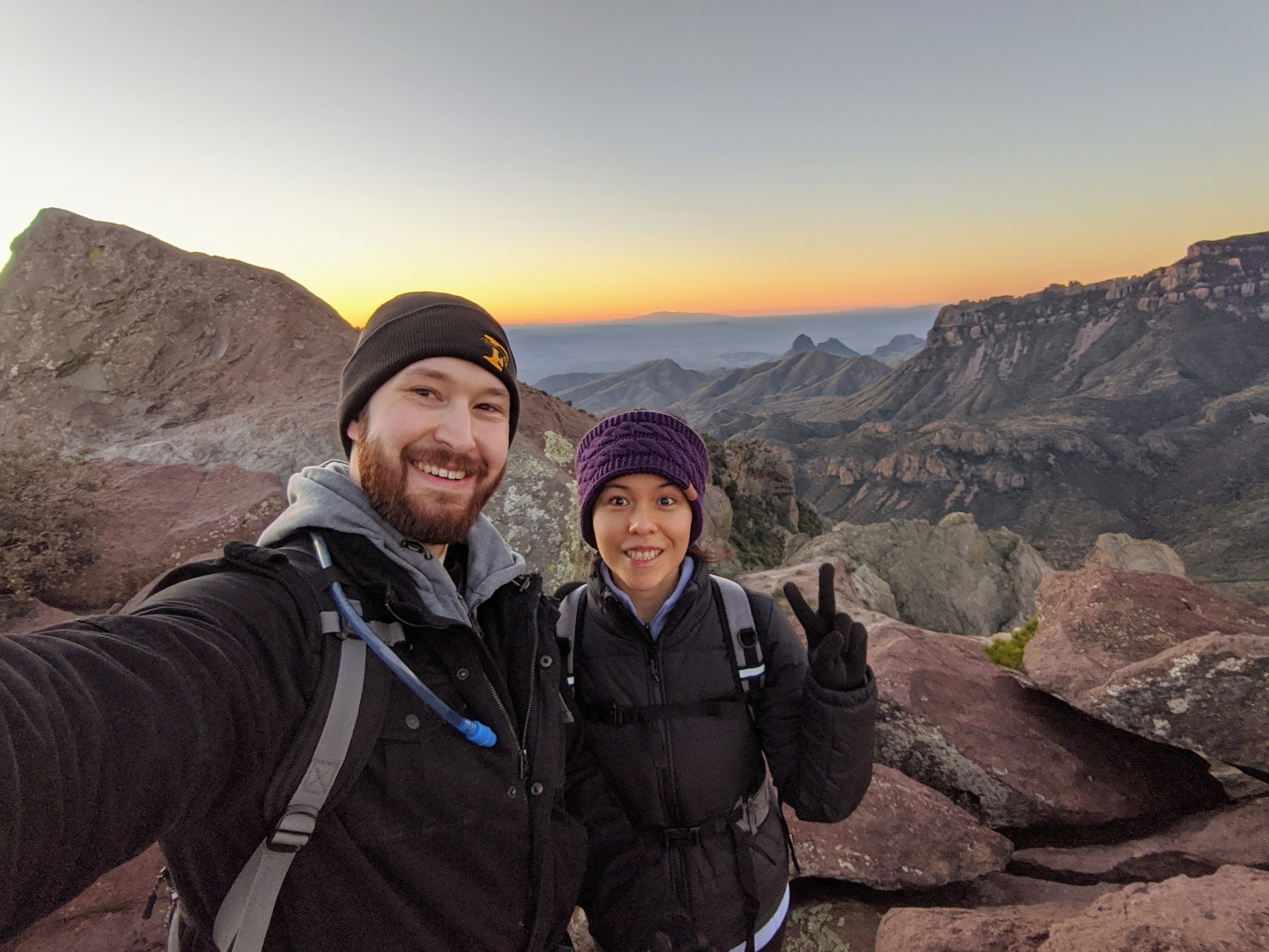 Leighton and his wife smiling at sunrise on a rocky summit in Big Bend, with canyon ridges and warm light stretching behind them.