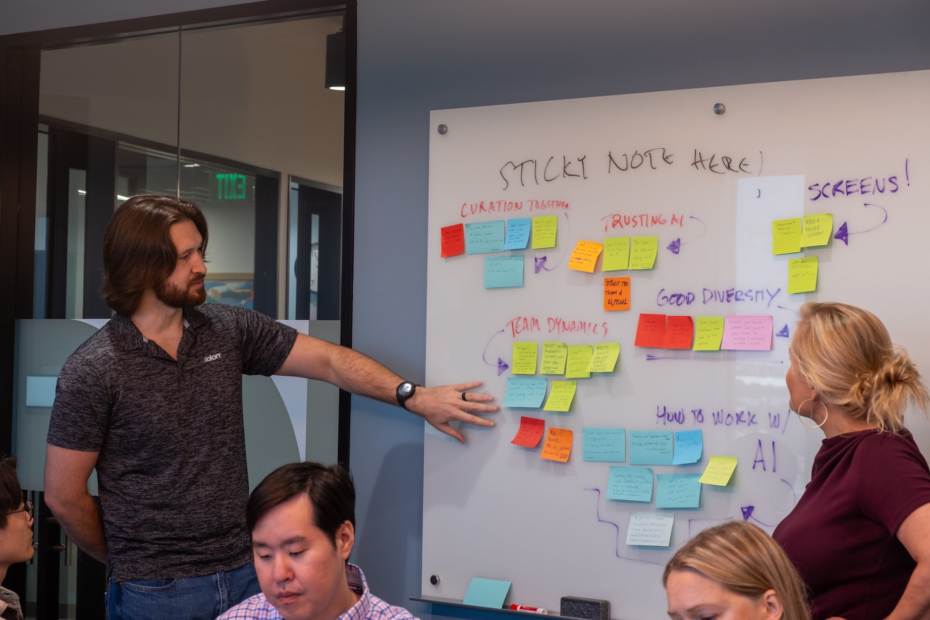 Leighton gesturing toward a whiteboard covered in sticky notes during a workshop. Handwritten section headers read Curation Together, Team Dynamics, Trusting AI, Good Diversity, Screens!, and How to Work with AI.