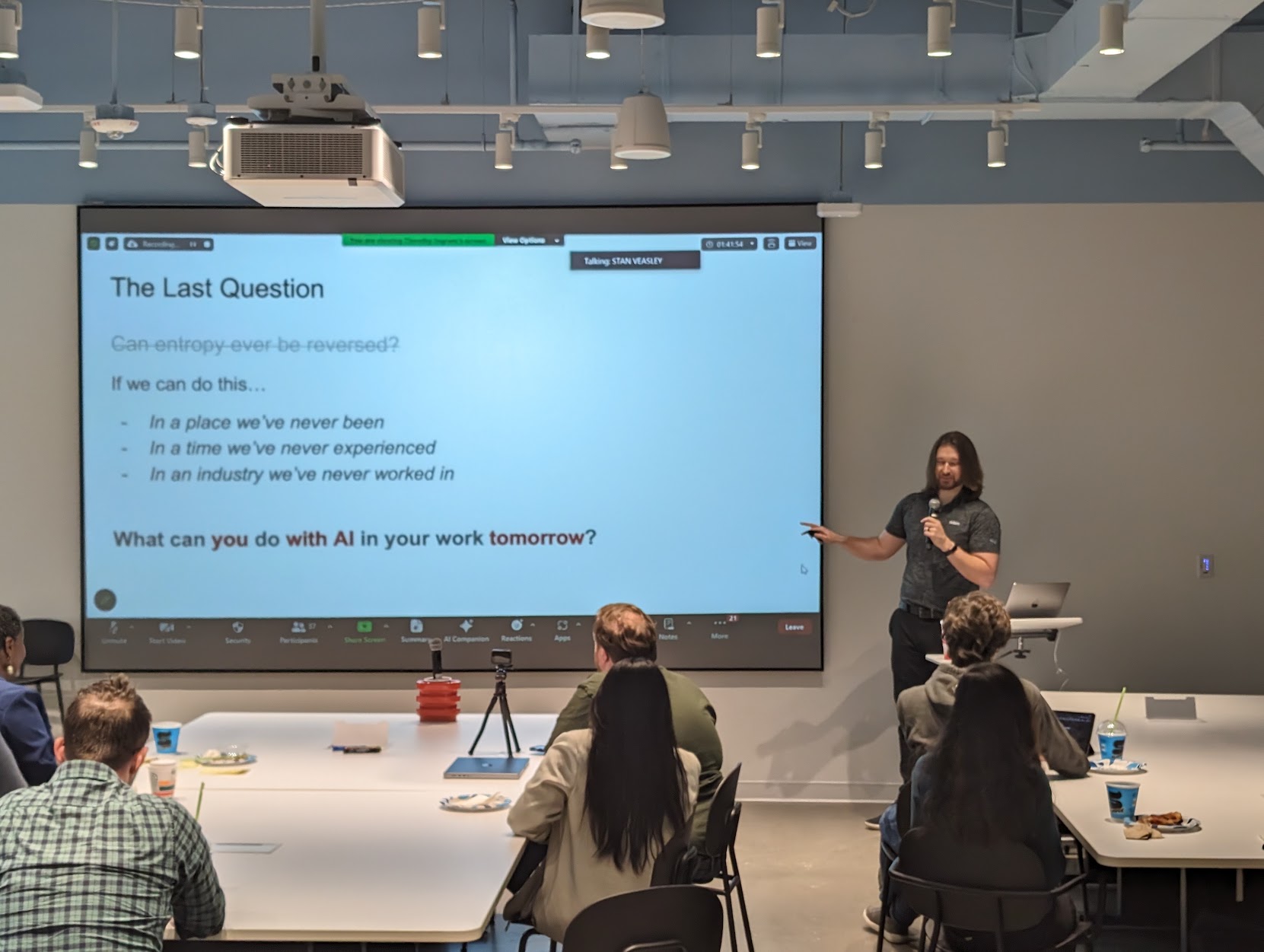 Leighton presenting at the 2024 SDN Dallas talk, gesturing toward a slide titled 'The Last Question' with the prompt 'What can you do with AI in your work tomorrow?'