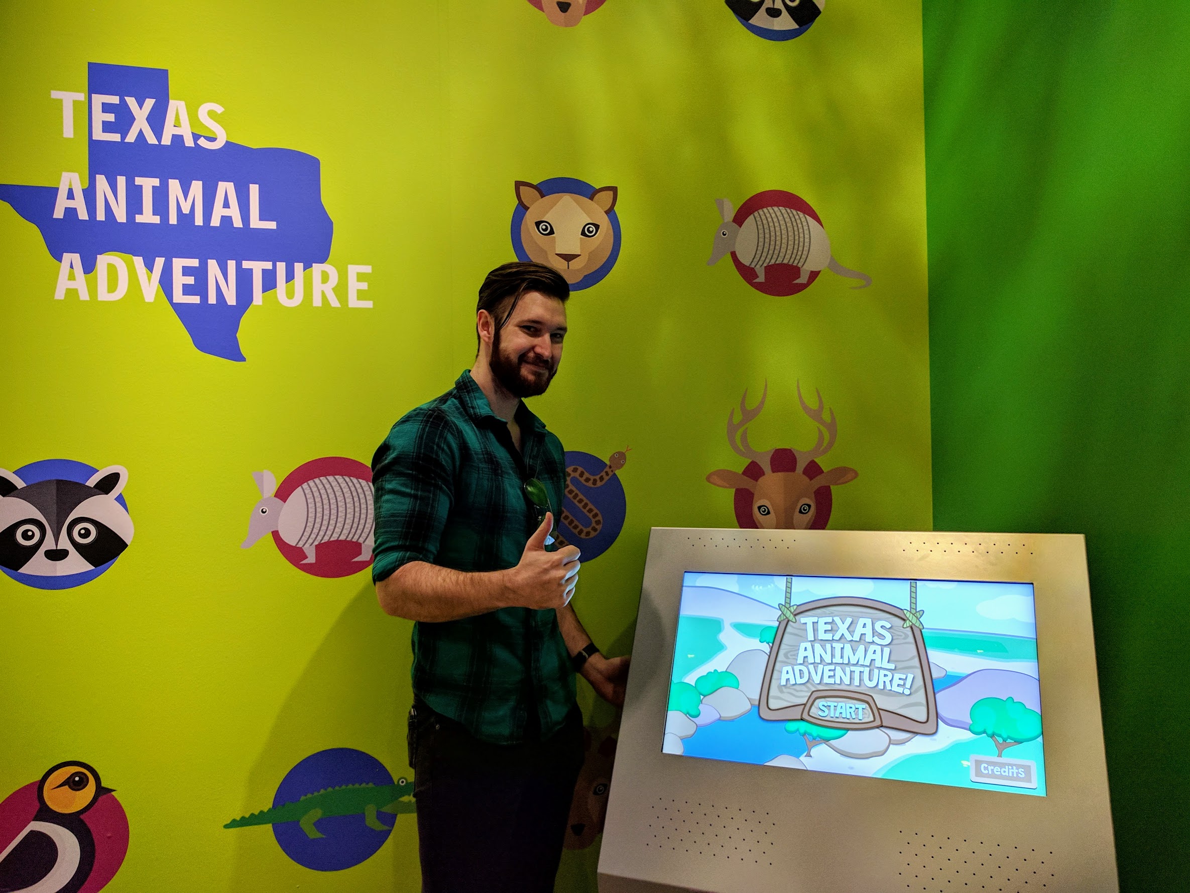 Leighton standing next to the Texas Animal Adventure kiosk in the Perot Museum of Nature and Science, giving a thumbs up in front of the game's title screen.